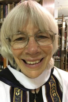 Close-up portrait of Rabbi Katy Allen, a modern Jewish American leader known for her environmental activism. She smiles warmly, wearing glasses and a traditional white clerical robe adorned with a detailed, colorful stole. Her friendly and approachable demeanor shines through, reflecting her commitment to faith and ecological sustainability. This image highlights her influence as a spiritual leader integrating religious values with environmental stewardship
