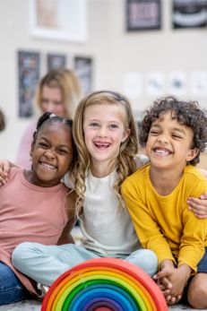 Three smiling children sitting together in a classroom with a colorful rainbow toy in the foreground.