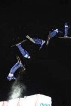 A sequence of images captures a skier performing an aerial stunt at night. The skier is dressed in a blue uniform and is shown in multiple positions as they execute a backflip, with snow spraying below them. The dark sky provides a stark contrast, highlighting the skier's movements and the dramatic nature of the jump.
