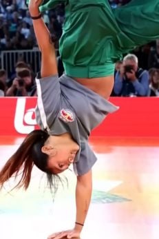 A breakdancer performs a one-handed handstand during a competition. The dancer is wearing a gray shirt with a logo patch and green pants, with long hair flowing down due to the inverted position. The background shows a crowd of spectators and photographers, with a red "lotto" banner prominently displayed behind them. The scene captures the energy and skill of the dancer in mid-performance.