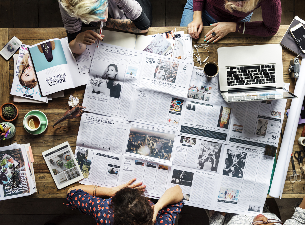  A group of people gathered around a wooden table, analyzing newspapers, magazines, and digital content on a laptop and tablet. The workspace is filled with coffee, notebooks, and creative tools, symbolizing media literacy, news analysis, and digital research.
