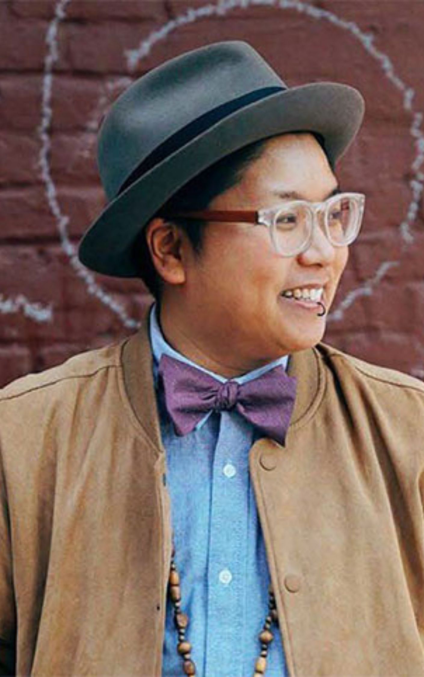 Kay Ulanday Barrett smiling while wearing a gray fedora, clear glasses, a purple bow tie, and a brown jacket; queer disabled poet and labor rights advocate.