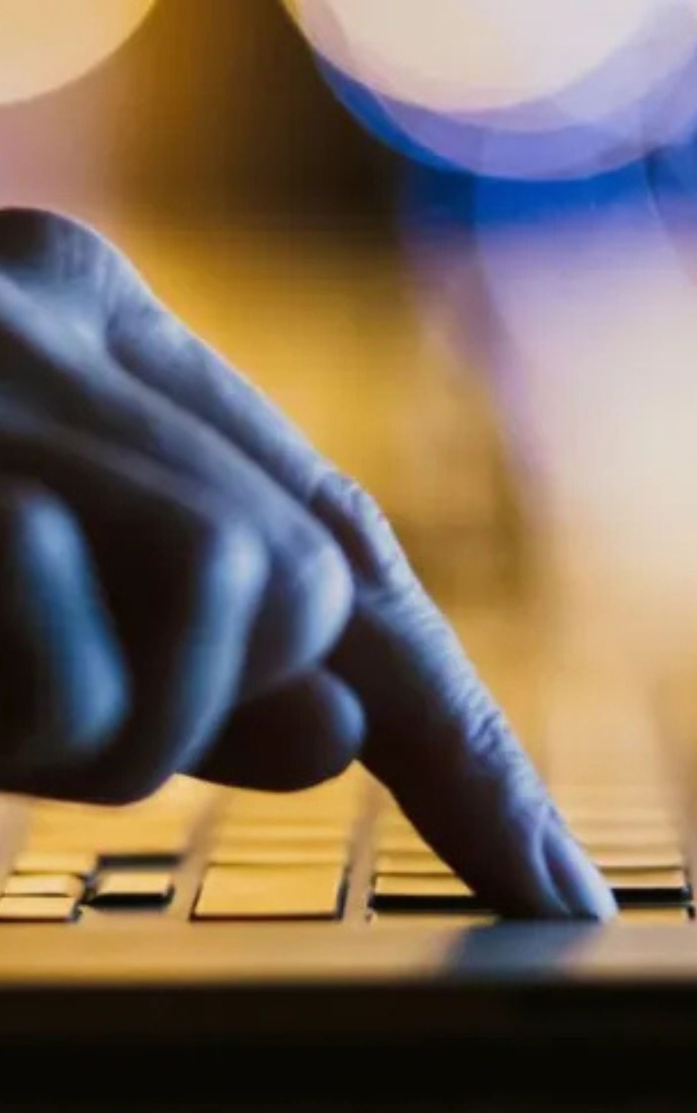 Close-up of a hand pressing a key on a laptop keyboard, with blurred background lights—symbolizing digital engagement, online activity, or media consumption.
