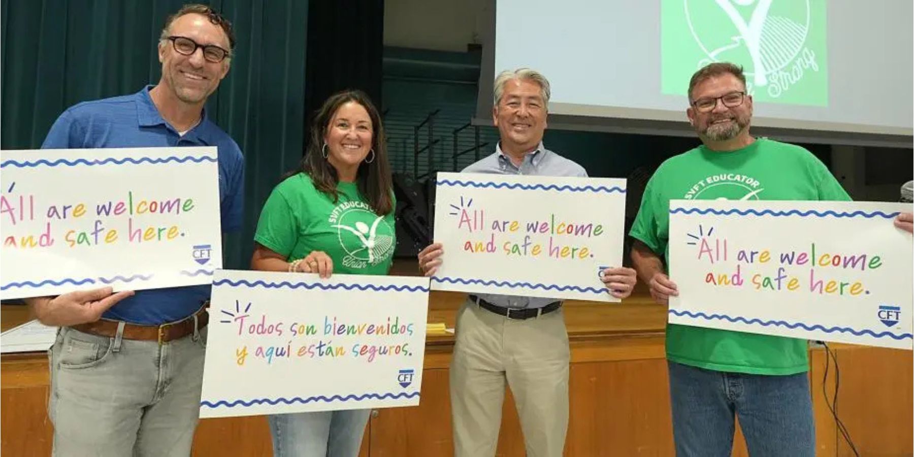 Four educators hold colorful signs reading “All are welcome and safe here” and “Todos son bienvenidos y aquí están seguros,” promoting inclusion and safety in schools.