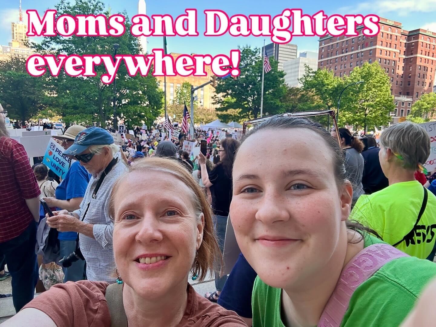 A smiling mother and daughter take a selfie at a No Kings Day rally, surrounded by a large crowd holding protest signs and American flags. The text “Moms and Daughters everywhere!” is overlaid in bright pink, reflecting the blog’s theme of intergenerational activism and the powerful role of women and families in democratic movements, as shared in Amber Chandler’s “No Kings Day: This Is What Democracy Looks Like.”