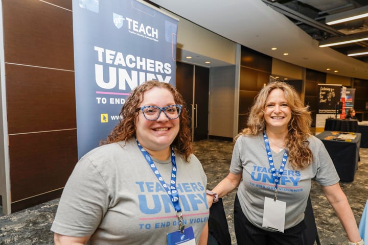 Sarah Lerner and Abbey Clements smile in front of a 'Teachers Unify to End Gun Violence' banner at the AFT TEACH 2025 conference. Both are wearing matching gray Teachers Unify t-shirts and conference lanyards.